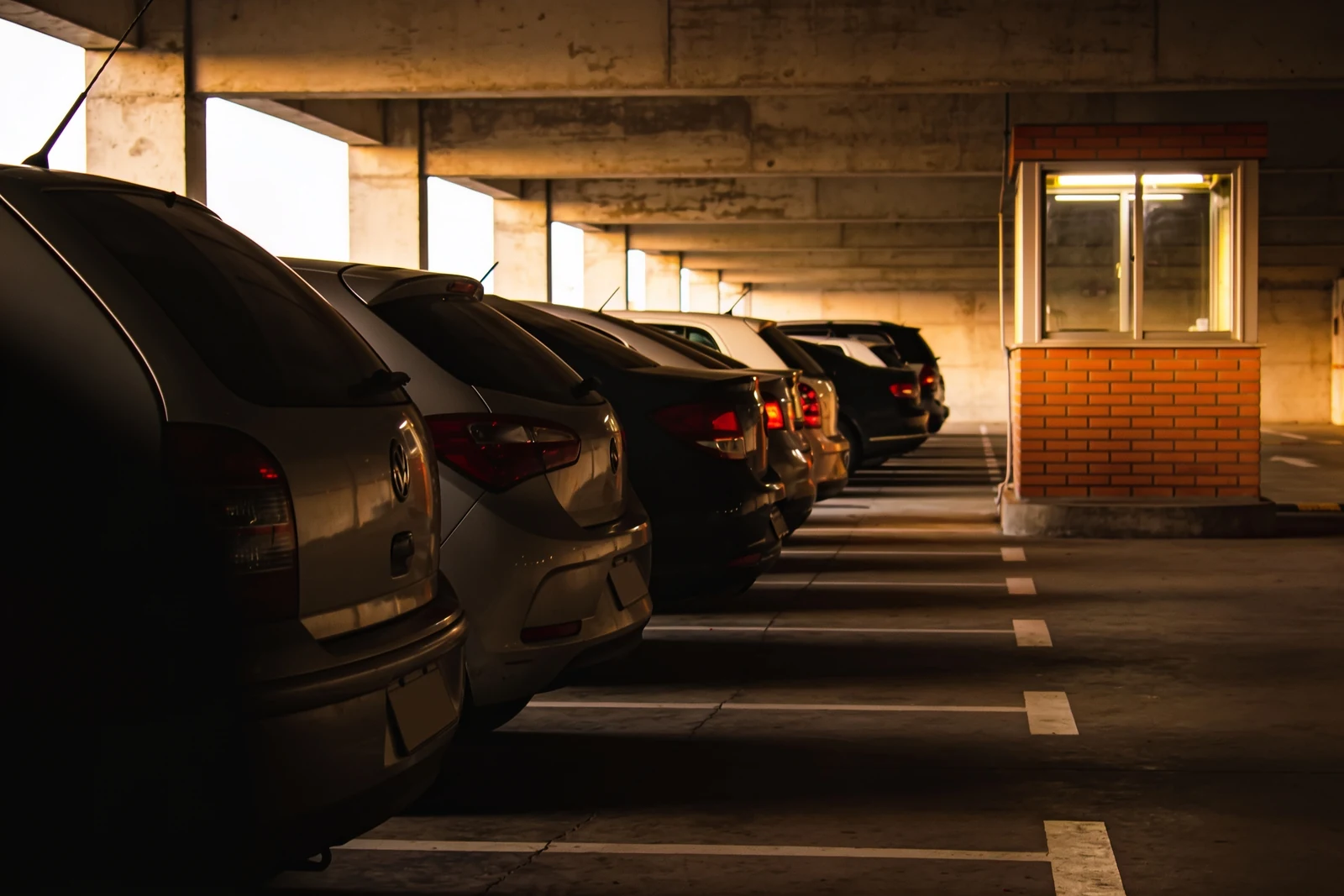 Estacionamento coberto em operação, com uma fileira de carros estacionados vista pela traseira e a guarita de controle iluminada ao fundo.